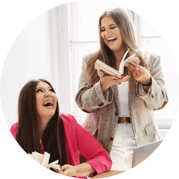 Na zdjęciu znajdują się właścicielki agencji kreatywnej Detalle - Beata i Ula. Kobiety trzymają w ręku otwarte książki i spoglądają się na siebie - są radosne i uśmiechnięte.